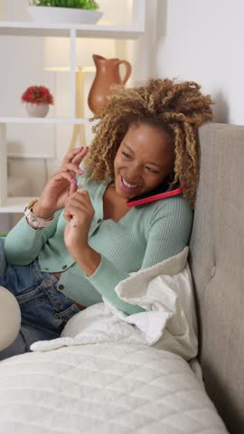 Young african american woman with curly hair talking on her smartphone while filing her nails. Relaxed, multitasking moment at home in her bedroom