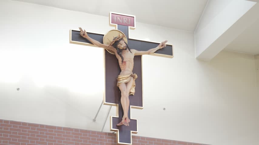 A wooden crucifix of Jesus Christ hanging on a white church wall above a brick pattern. Low angle static shot