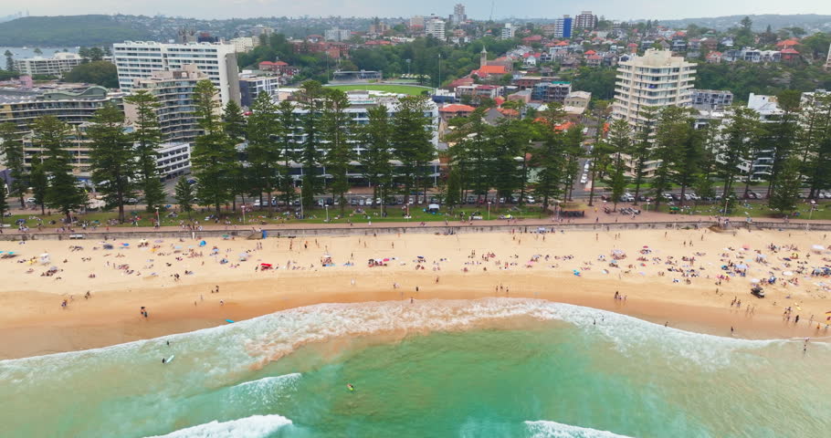 Aerial view of Manly Beach in Sydney, Australia, turquoise ocean waves breaking on the sandy shore, people relaxing, swimming, and surfing n summer weekend. Coastal buildings houses line the promenade