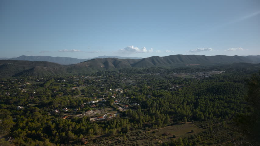 Elevated rural valley panorama with scattered homes, wooded slopes, farmland patches, and distant mountain ridges under clear blue sky