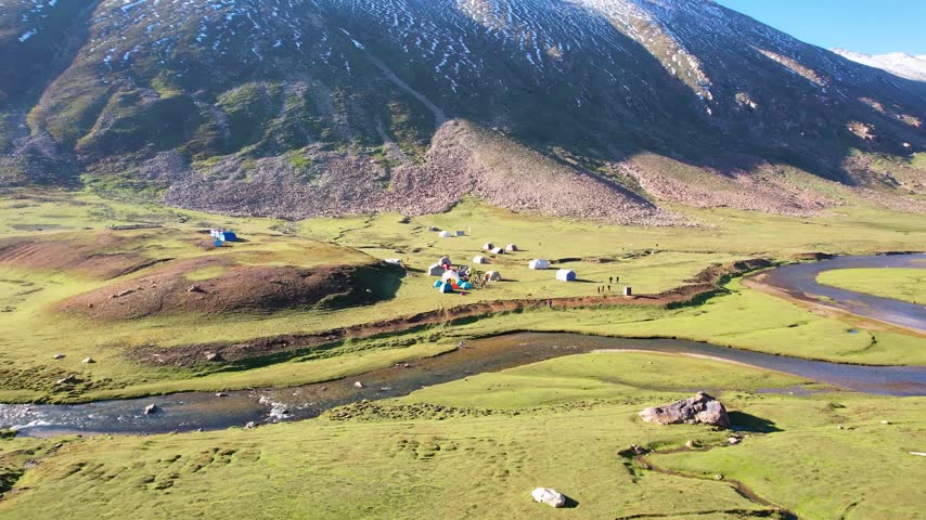 High-angle, wide aerial shot overlooking an outdoor expedition base camp setup in a sprawling alpine meadow valley. The footage captures a collection of colorful dome and frame tents clustered along a clear, winding mountain stream, bordered by rolling green hills and a steep rocky mountain slope in bright morning sunlight.
