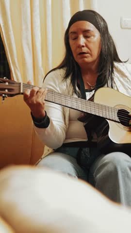 Focused mature woman with long hair sitting on a couch and passionately playing a classical acoustic guitar, practicing chords and melody in a cozy living room during her leisure time