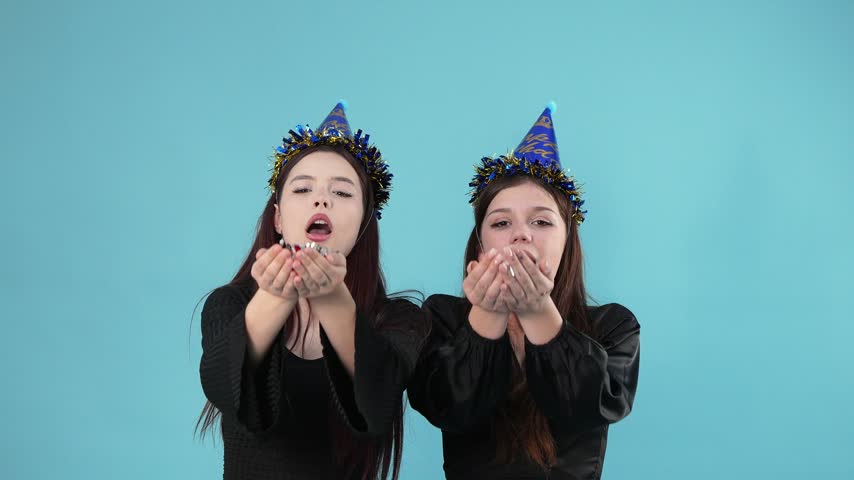 Two joyful young women in party hats blow star-shaped confetti at the camera against a blue background, then jump up and down happily and embrace in a warm hug.