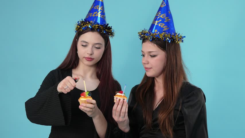 Two young women wearing blue party hats stand against a cyan background. One girl lights a candle on a cupcake using a lighter, sharing the flame with the other cupcake.
