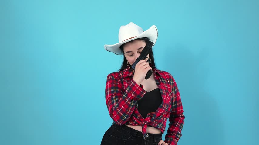 A young woman wearing a white cowboy hat and red plaid shirt holds a black gun, smiles, winks at the camera, and turns sideways against a bright blue studio background.