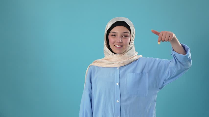 A smiling young Muslim woman wearing a beige hijab and light blue shirt points downward against a solid blue studio background, indicating a link or call to action below.