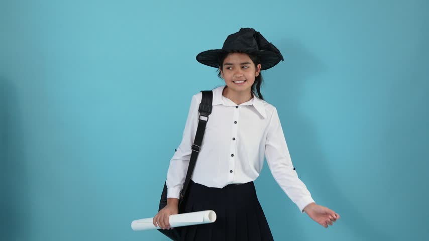 A young girl in a white shirt and black witch hat poses against a blue background. She takes off her hat, puts it back on, holds rolled paper scrolls, and smiles playfully at the camera.
