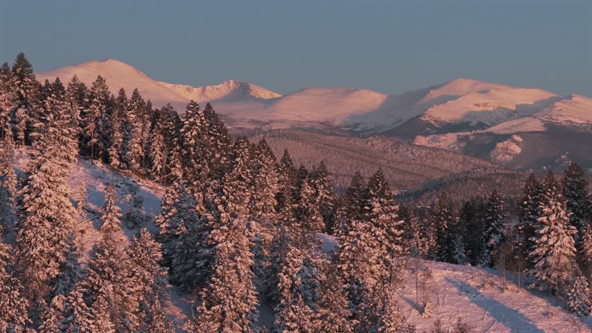Dramatic sunset light illuminating snow covered pine forest with massive rocky mountain peaks rising in distance under clear winter sky, slow panoramic aerial perspective.