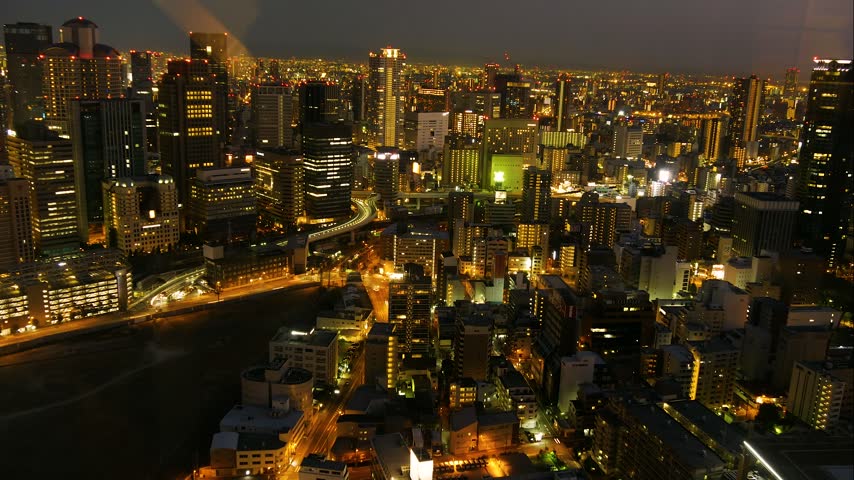 High angle aerial footage of vibrant Japanese metropolis at night showcasing dense urban architecture bright city lights moving traffic and cinematic atmosphere in Osaka Japan.