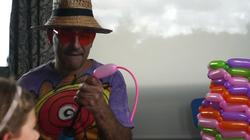 A male entertainer wearing a straw hat and glasses inflating a long pink twisting balloon with a manual air pump in a bright room.