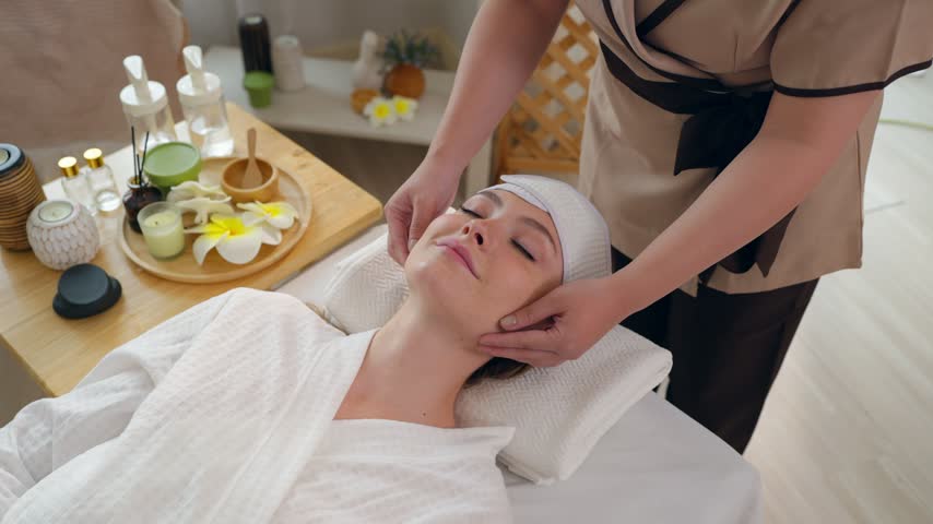 Beautiful woman getting head and facial massage treatment at beauty spa. Attractive girl lying down on massage bed, getting relaxing physiotherapy service from masseuse for body care in spa clinic.