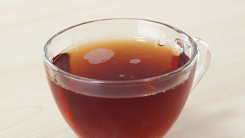 White granulated sugar is poured into a clear glass cup filled with hot black tea.