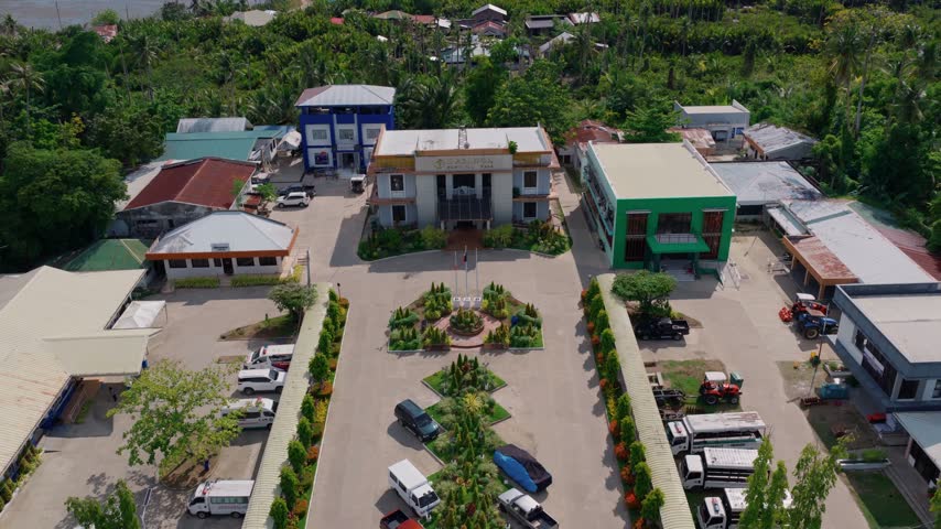 Aerial drone footage showcasing municipal hall complex in Philippines with parking lot surrounded by tropical vegetation trees and nearby residential houses during sunny day.