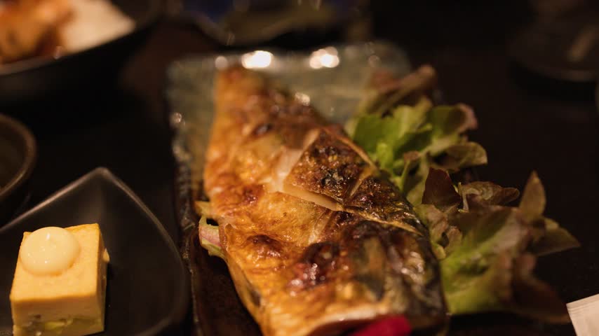 Chopsticks pick apart a grilled mackerel fillet served with sides in a restaurant.
