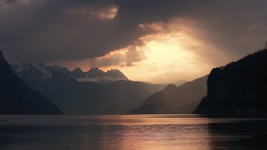 Cinematic view of golden sunset rays filtering through heavy dark storm clouds above tranquil mountain lake waters with silhouette trees in foreground during moody evening dusk.