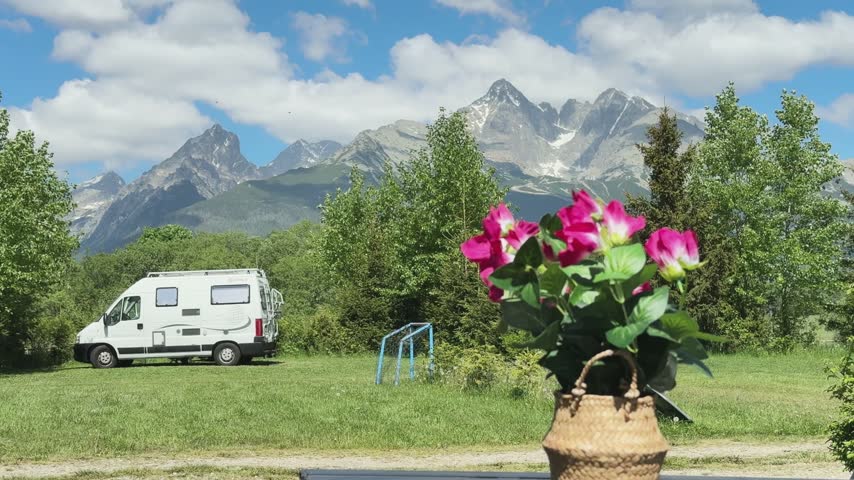 Scenic view of recreational vehicle parked on green meadow featuring basket of bright pink flowers in foreground under majestic jagged mountain peaks and blue sky with white fluffy clouds.