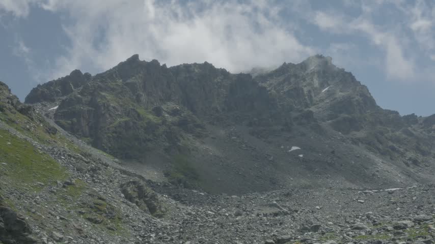 Steep craggy mountain summits dominate dramatic landscape with drifting clouds and barren rock scree fields. High altitude terrain captures powerful natural geological beauty.