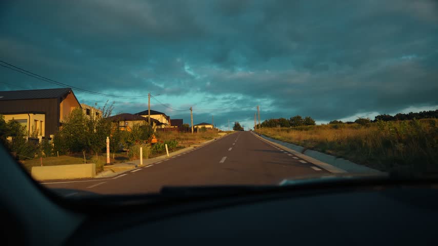 A quiet rural road leading through a residential area under a dramatic stormy evening sky, with houses, utility poles and open fields.