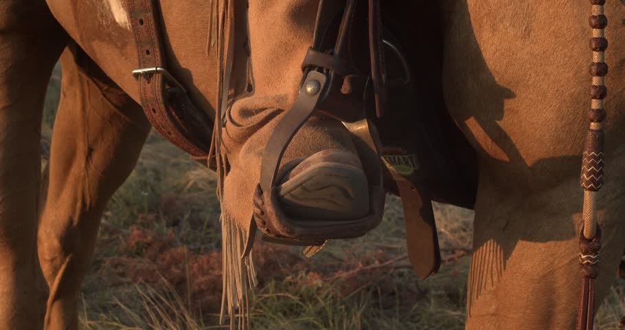 Cowboy boot rests in a leather stirrup on a saddled horse at warm sunset, emphasizing classic western ranch life.