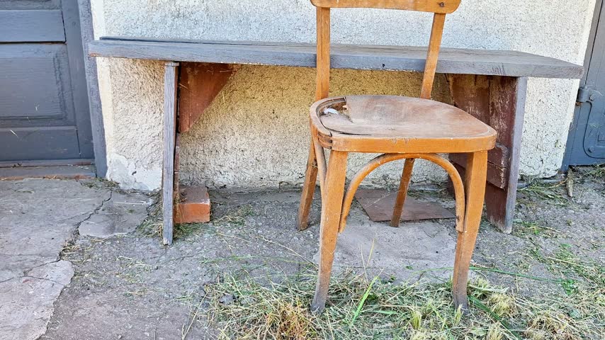Wooden chair and simple bench standing outside an old house, with weathered walls, cracked pavement, and a rustic exterior creating an authentic countryside setting