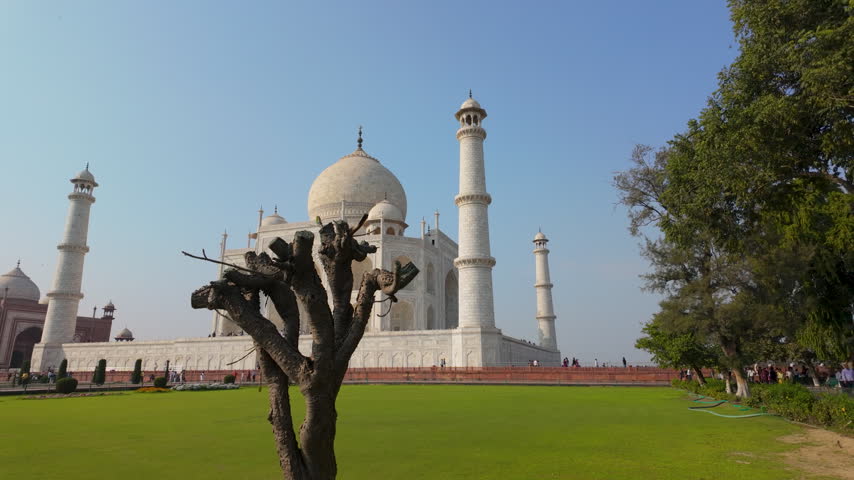 A wide shot of the iconic Taj Mahal mausoleum in Agra, India, featuring the white marble architecture, lush green lawns, and a clear blue sky. Suitable for travel and history documentaries.