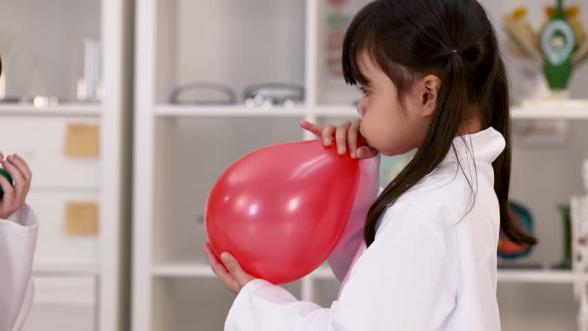 A young girl in a white lab coat inflates a red balloon in a classroom.