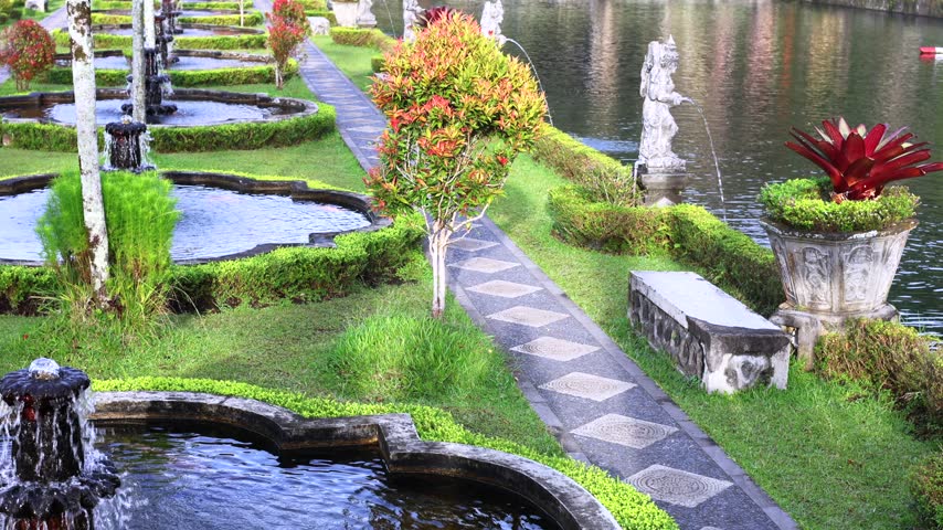 Ornamental garden fountains shimmer beside quiet pond in sunny tropical park. Curved water basins, stone statue, trimmed lawns and patterned walkway create serene landscape with vivid greenery.