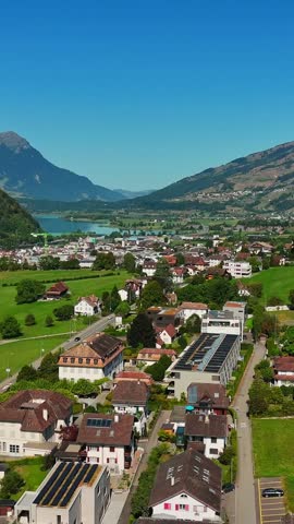 Vertical aerial view over Schwyz, Switzerland, showing residential homes, rooftop solar panels, green meadows and Lauerzersee beneath steep Alpine slopes.