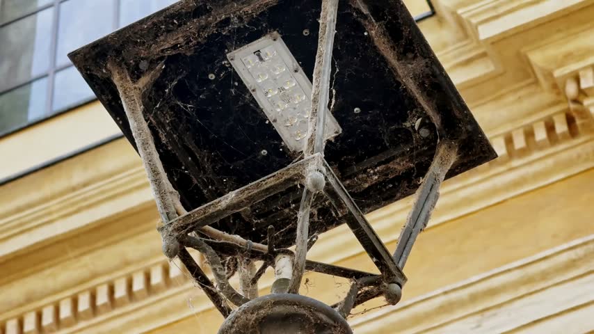 Close-up of an old street lantern covered in cobwebs, revealing a modern LED module against a historic yellow building in Parma, Italy.