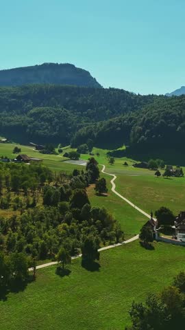 Vertical aerial view of Schwyz, Switzerland, showing a winding country path through green meadows, scattered homes and forested Alpine hills beneath a clear summer sky.