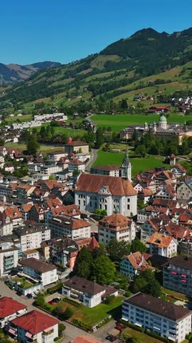 Vertical aerial view over Schwyz, Switzerland, centered on St. Martin Church and the historic town core, with red-tiled roofs, green fields and steep Alpine slopes beyond.