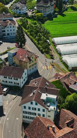 Vertical aerial view of Schwyz, Switzerland, with cyclists and cars traveling past homes, greenhouses and cultivated fields in a sunny Alpine town.