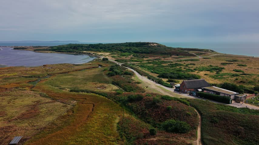 Aerial footage across Hengistbury Head in Dorset, England, showing heathland, coastal wetlands, woodland and walking trails beside Christchurch Harbour. Visitor facilities sit alongside the main route