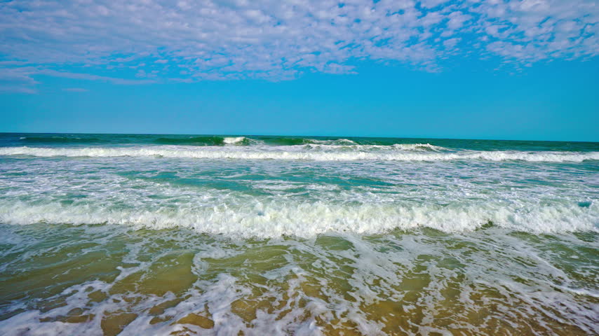 Sea beach background with blue summer sky. Tropical ocean waves over sand near shore. Dreamy sea beach on tropical coast. Seaside tranquility beside open ocean water.