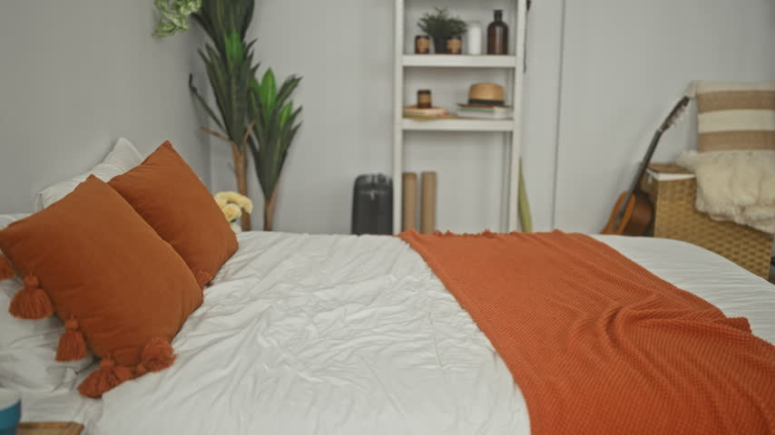Man points remote control with hand while seated on bed in building, orange pillows and blanket visible; relaxed leisure comfort.