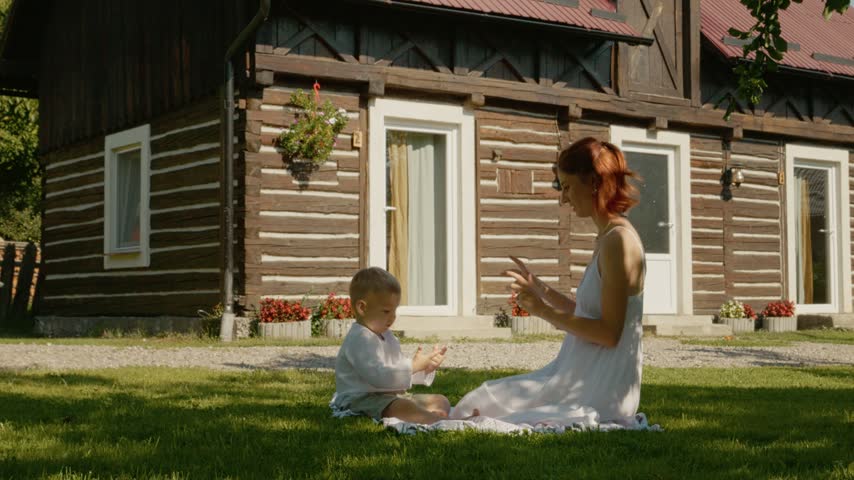 A mother and her child sit on the grass. They are playing and enjoying activities in front of a traditional house located in Bucovina. The sun shines on them as they laugh.