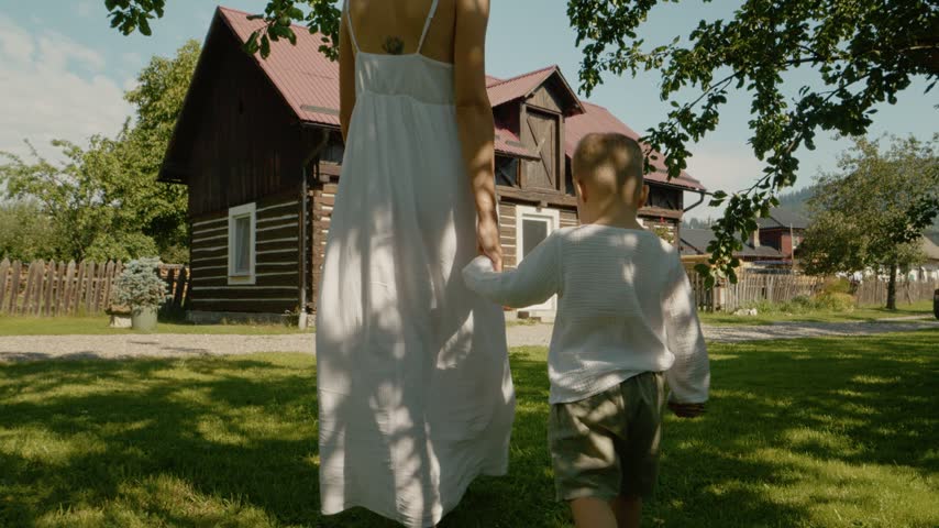 A woman and her boy stroll hand in hand past a traditional house in Bucovina. The scene captures a sunny day with greenery around them and a glimpse of the house.