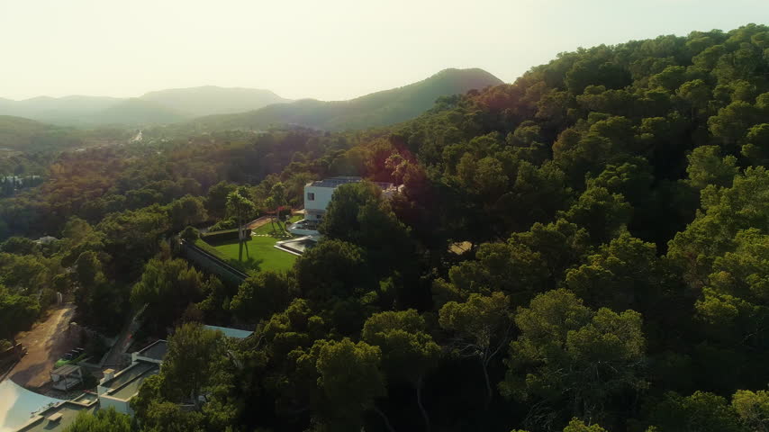 Drone retreats from a hillside home through warm rays over Mallorca forest.
