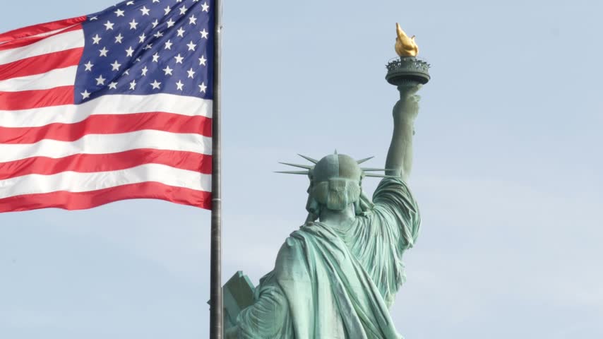Statue of Liberty on Liberty Island, New York, United States. Cooper monument from behind, democracy and patriotism symbol near Manhattan, NYC USA. American flag waving in wind. Independence concept.
