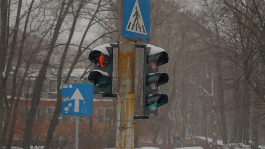Snow continuously falls over urban streets, creating a winter wonderland. A traffic light, partially covered in white, signals the busy streets below.
