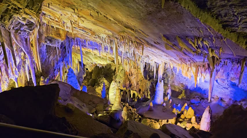 Ancient cavern ceiling covered in delicate stalactites and mineral deposits, illuminated by spotlights that reveal layered limestone textures, shadows, and natural underground formations.