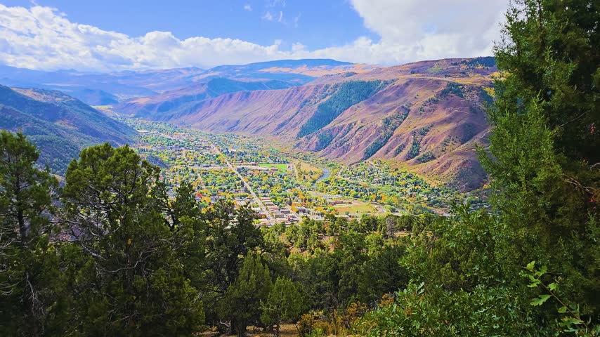 Scenic viewpoint above Glenwood Springs, Colorado, revealing neighborhoods, the Colorado River, evergreen forest, golden fall foliage, and rugged Rocky Mountain terrain in clear daylight.