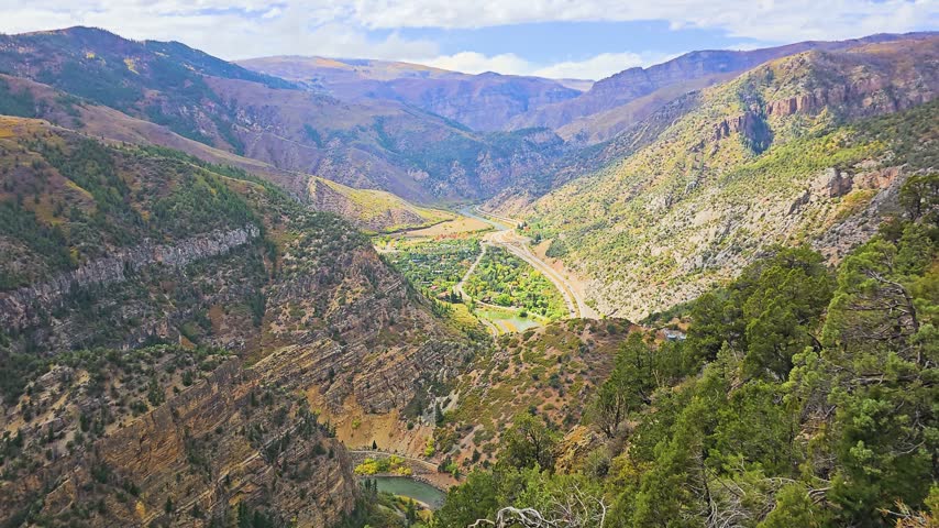 Sunlit overlook of Glenwood Springs, Colorado, with layered mountain ridges, dense green forest, golden fall foliage, the Colorado River, valley homes, and distant peaks beneath clouds.