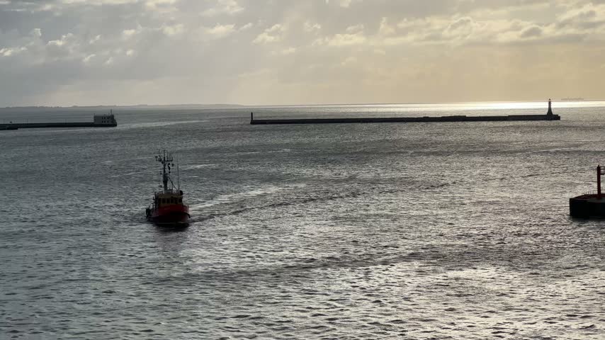 Seaport entrance with moles and berths against the cloudy sky and the water surface with light waves and ripples, view close-up from the vessel entering the port at sunrise while panning
