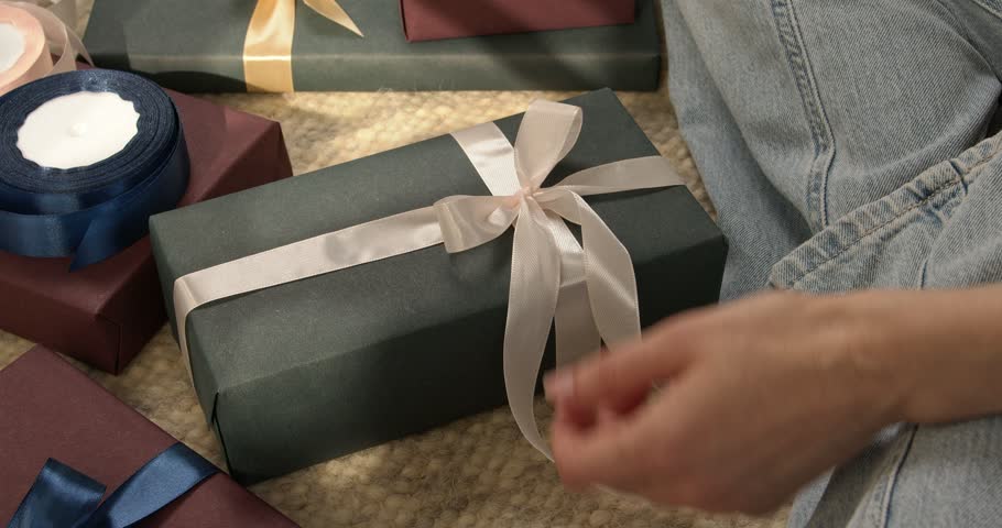 Woman hands tying white satin ribbon on gift box with other presents around close up video.