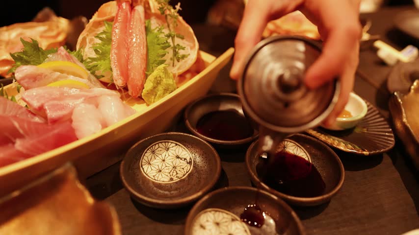 Close-up shot of someone adding soy sauce to dishes. A boat filled with fresh raw fish sashimi is visible.
