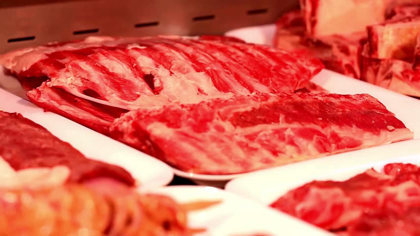 Fresh raw beef ribs and various cuts of red meat arranged on white trays inside refrigerated retail display case ready for sale at local butcher shop.