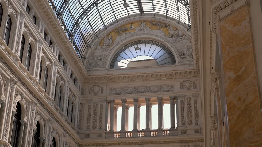 Ornate interior facade and arched glass roof of Galleria Umberto I in Naples seen in a steady upward view.
