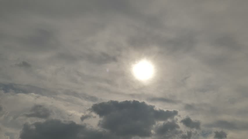 A bright white sun shines through a layer of thin grey clouds in an overcast sky. Darker, denser cloud formations drift slowly across the lower portion of the frame, creating a calm, layered atmosphere. Occasional birds fly across the scene, adding subtle movement to this softly lit, diffused-light sky. The clip offers a serene, muted aerial view with gentle cloud motion and no audio.