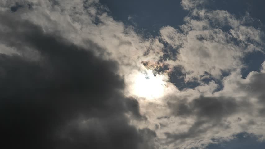 Bright sunlight shines through a layer of moving clouds in a wide blue sky. Dark grey clouds drift steadily across the frame from left to right, intermittently obscuring and revealing the sun and creating shifting backlighting. Small dark birds fly quickly through the frame at various points during the clip. No audio.